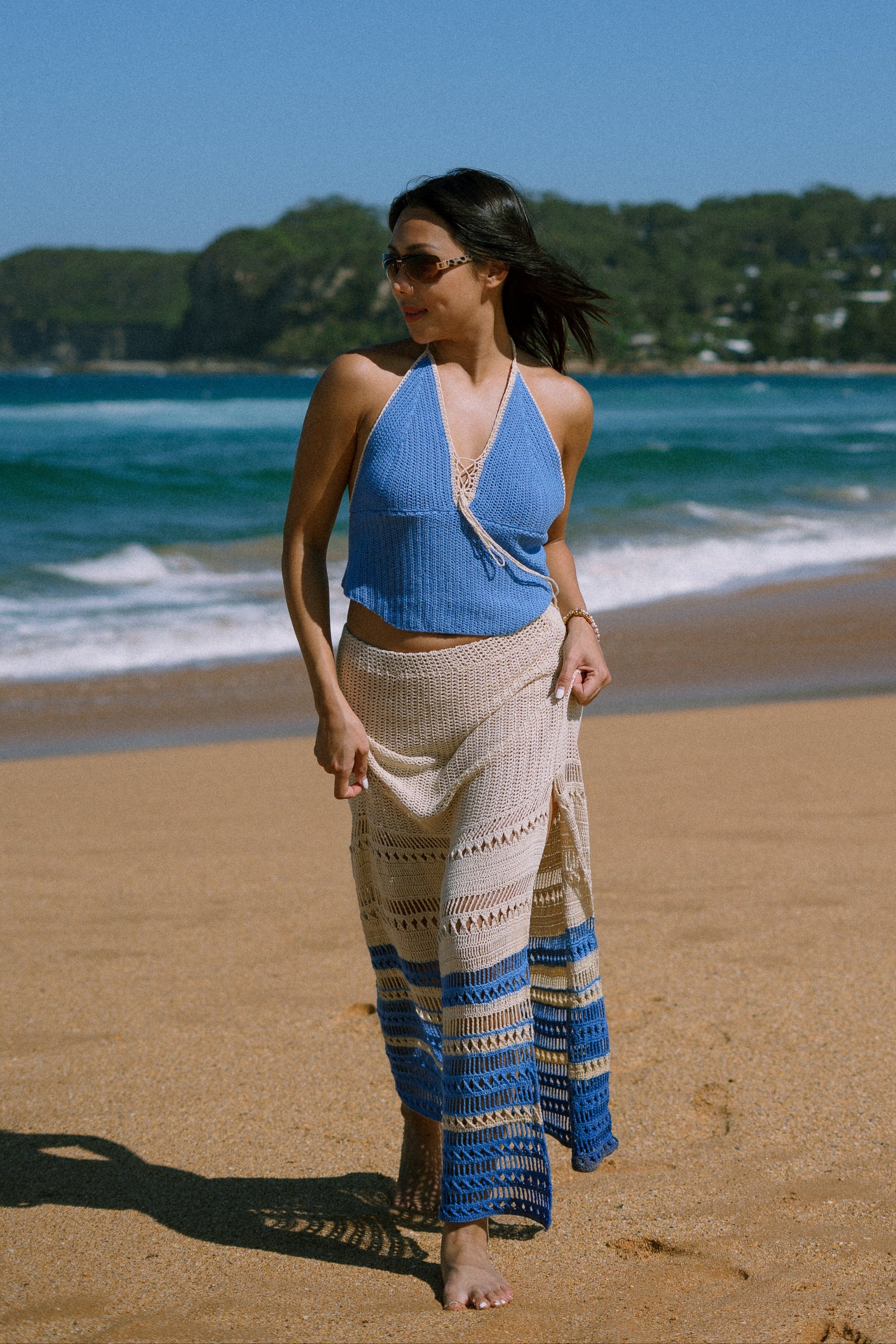 Woman in a blue crochet top and crochet patterned skirt standing on a beach with ocean and trees in the background