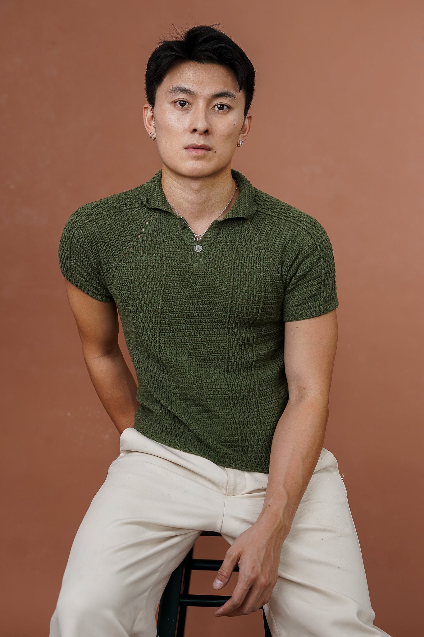 Man wearing a green crochet polo shirt and white pants against a brown background