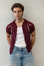Man wearing a maroon crochet shirt over a white tank top and blue jeans against a plain background