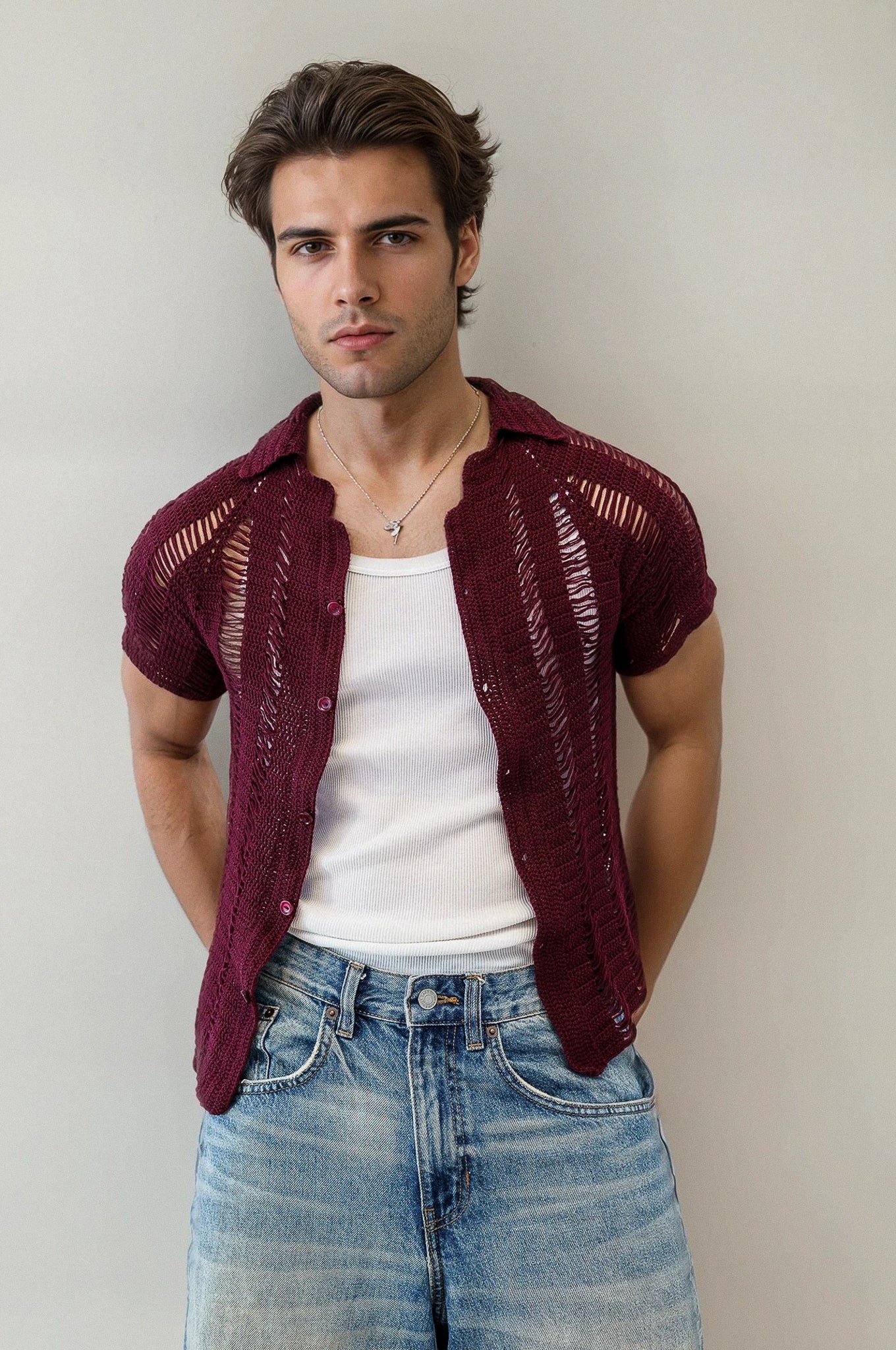 Man wearing a maroon crochet shirt over a white tank top and blue jeans against a plain background
