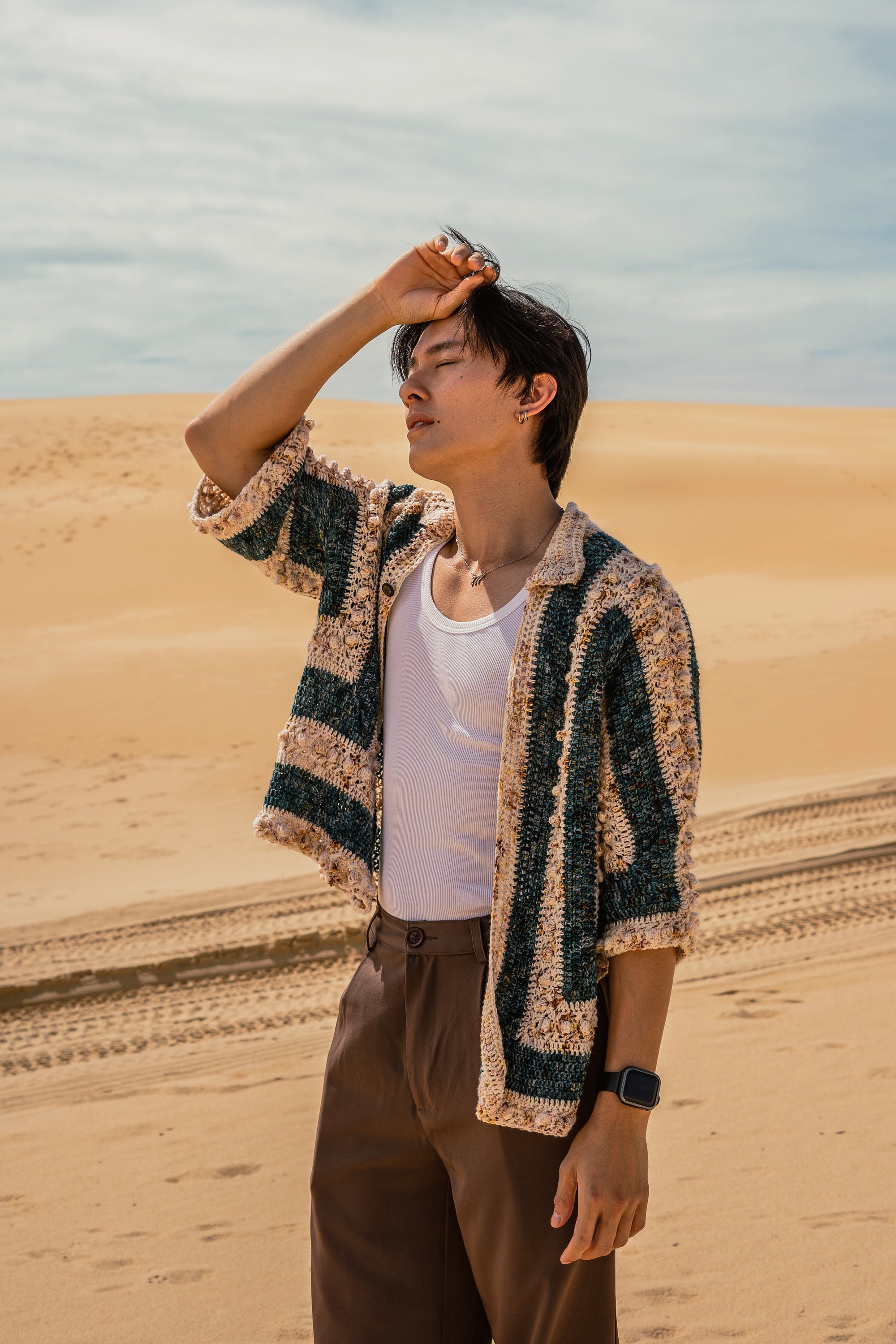 Person standing in a desert with sand dunes and a clear sky, wearing a striped crochet pull over